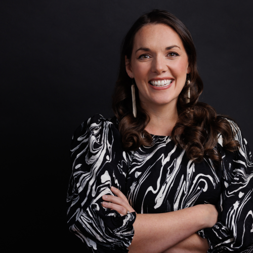 a white woman with shoulder length brown hair stands with her arms folded. she smiles and wears a black and white top.