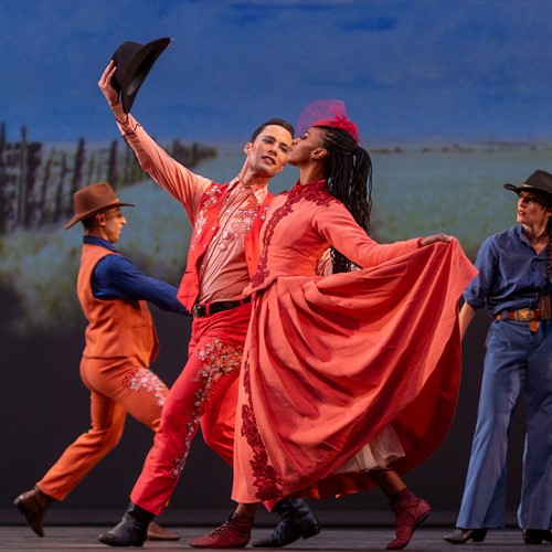 in a dance performance, a black woman in a full-skirted red western-style dress walks alongside a white man dressed as a cowboy in a red outfit