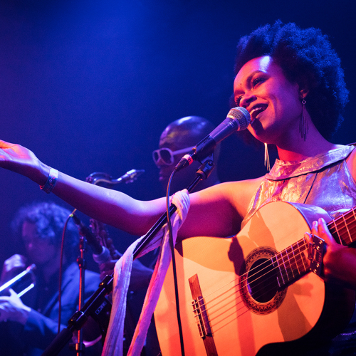 an ethiopian-american woman in a flowing golden top extends an arm as she sings on stage