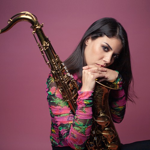 Melissa Aldana, a woman with brown hair sits holding a saxophone with her hands at her chin in front of a pink background.