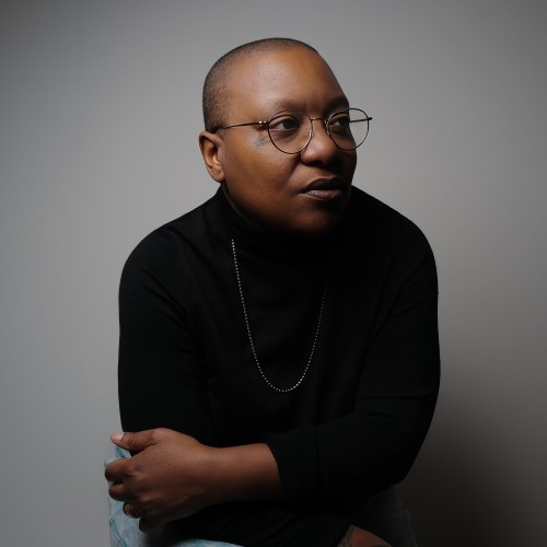 a black woman with a shaved head and thin framed round glasses. She sits with her arms around herself, leaning slightly forward, looking at us from the side