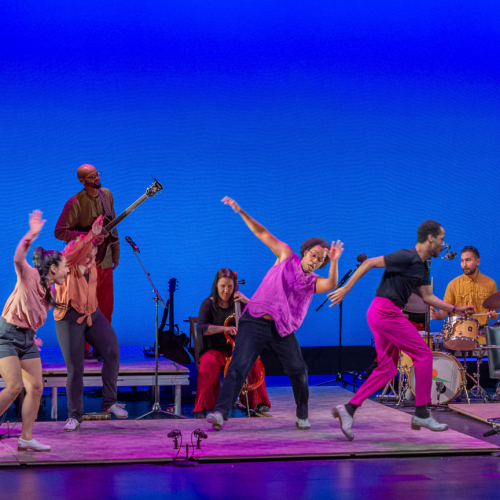 A multicultural group of tap dancers and musicians perform on low platforms