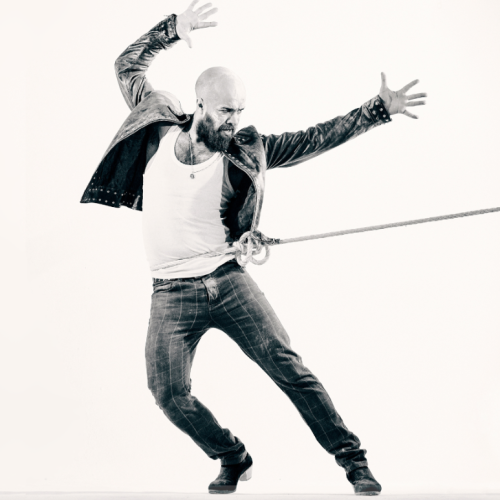 a bald-headed spanish man in heeled boots, jeans, and a leather jacket is tied with a rope around his waist. he leans away from the rope, arms raised