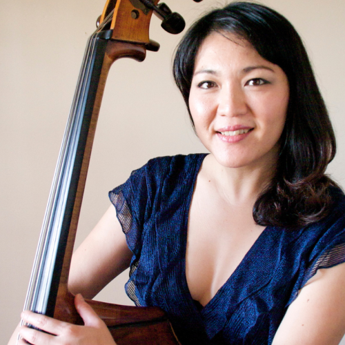 a long-haired japanese woman in a cap-sleeved dark blue dress. she stands with an arm around her double bass and smiles