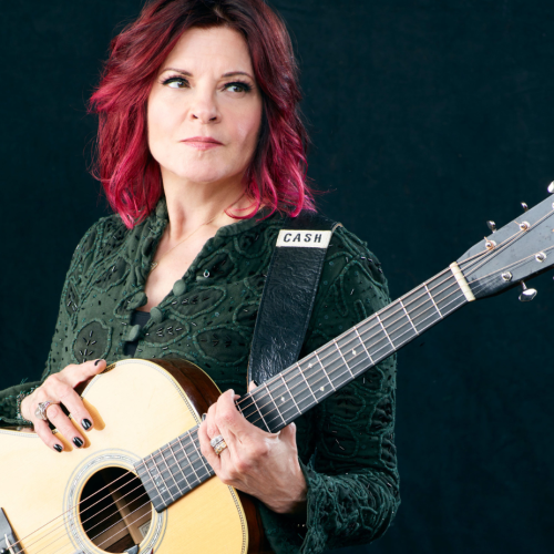 a white woman with vivid burgundy hair holds a guitar that reads CASH on the strap. She looks away out over the headstock of the guitar.