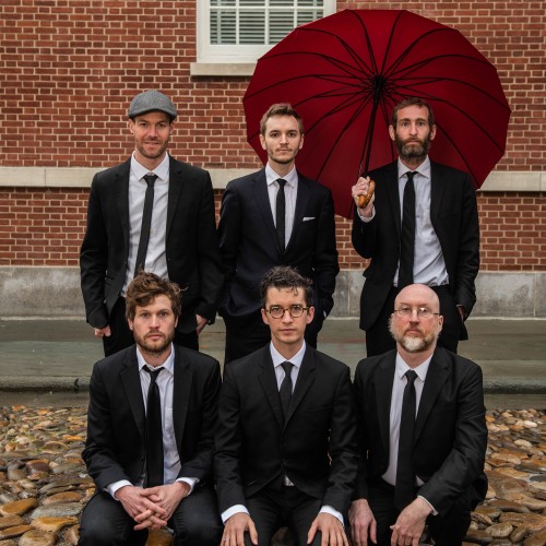 six white men of varying ages in suits, one with a red umbrella, in front of a brick building