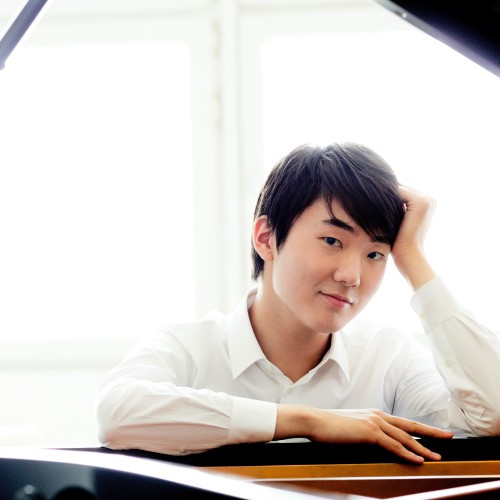 An Asian man with dark hair in a white shirt sits at a piano in front of a whitewashed window. 