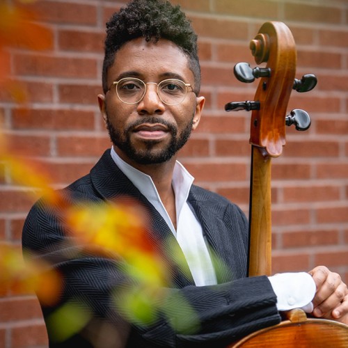 with an out-of-focus branch in the foreground, a young black man with wire-rimmed and a short beard wraps his arms around his cello