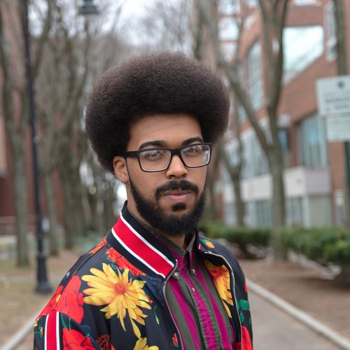 a puerto rican-dominican man with black glasses and a vivid floral athletic-style jacket. he is outdoors, on a brick walkway, on a college campus or similar location.