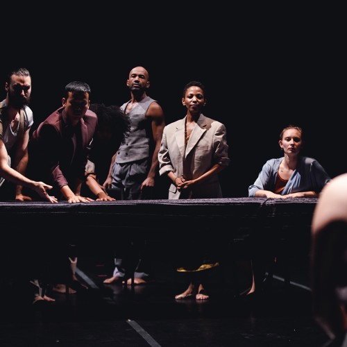 a diverse group of five men and women lean over a long table, as if challenging a person whose back is turned to us