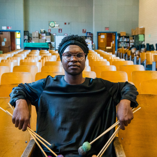 a nonbinary black person holds two sets of percussion mallets in their hands. they sit in a public school auditorium, with their elbows extended over wood-backed seats