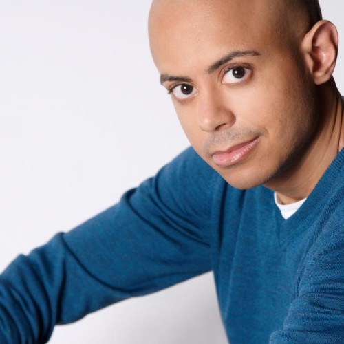 a man with light brown skin and a bald head sits with an arm draped over his knee. he wears a deep blue lightweight sweater