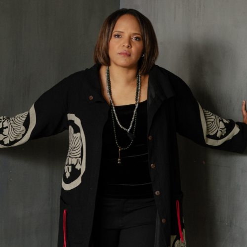 a Black woman in a black outfit topped by a long draped black jacket with abstract ivory medallion designs stands in a corner, with her hands on the concrete walls to either side of her.