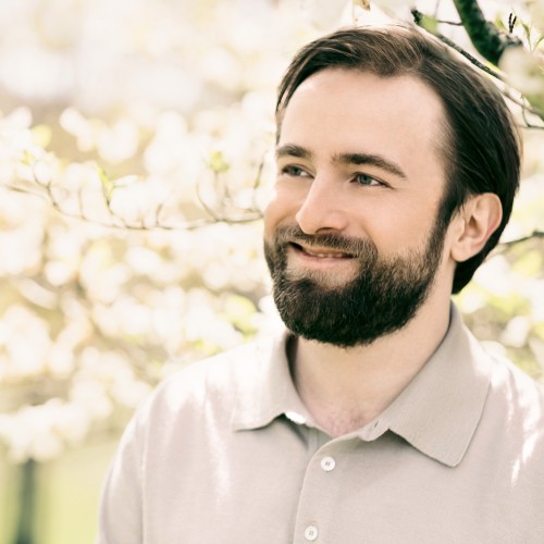 a bearded man smiles, he stands under a flowering tree