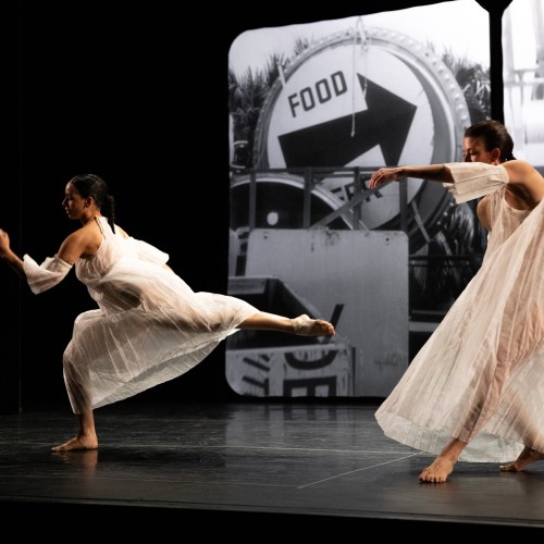 in front of a black and white photograph of roadway signs, two female dancers in sheer flowing ivory dresses move across the stage, one leaning forward on a bent leg, and the other with an arched back