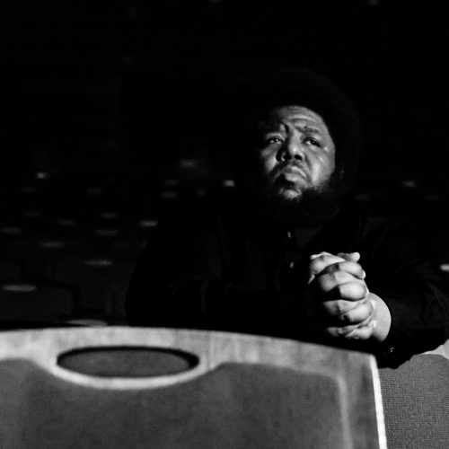 a low-contrast image of a black man in theater seating, his hands clasped and wrists resting on the seatback in front of him
