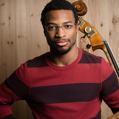 a black man in his late 20s, with glasses and short hair and wearing a red striped sweater, stands in front of a wood-paneled wall with his bass resting against his left shoulder. 