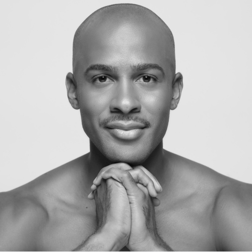 a bald headed black man with a mustache clasps his hands below his chin. he is shirtless, and he has a slight smile as he gazes serenely at the camera