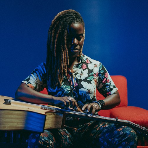 a young Black woman with shoulder-length locs leans over her guitar, which she holds in her lap.