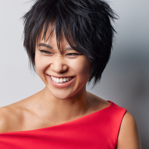 a glamorous chinese woman in her early 30s wrinkles her nose as she laughs. Her hair is in a chopped shag style, and she wears a red one-shoulder dress