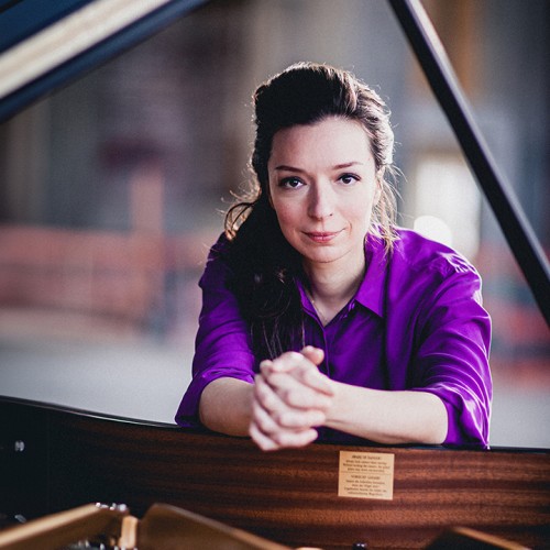 a dark-haired russian woman in her early 30s wearing a rich purple shirt leans over the edge of an open grand piano