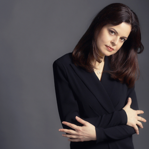 a dark haired white woman inclines her head, her arms folded across her chest. She wears a black blazer