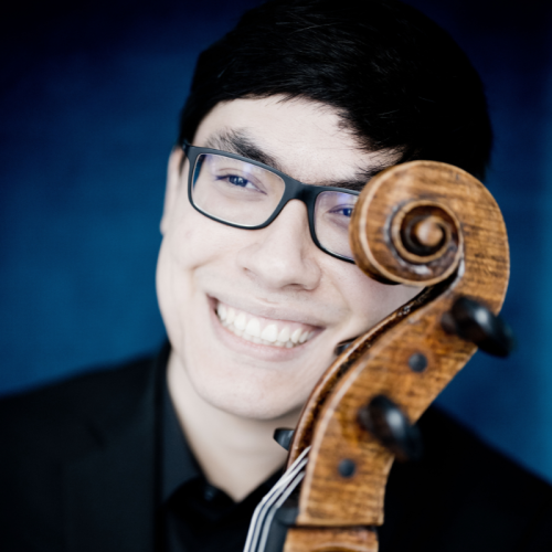 a man in his 20s, of eastern european and chinese heritage, smiles with the scroll of his cello curving across one cheek, partly covering his eye and eyeglasses