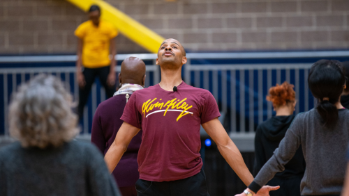 a bald black man in his 30s leads a class in a breath exercise. his arms are extended down at his sides, and he lifts his face looking over the participants