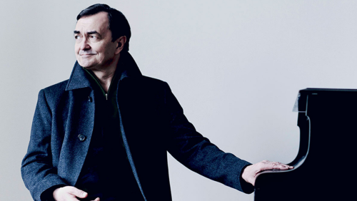 pierre-laurent aimard rests one hand on a grand piano. an inset image of a book with a historical drawing on the cover