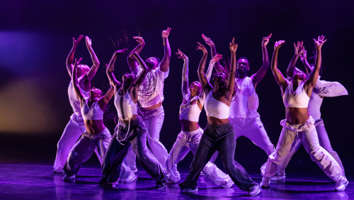 a row of dancers in white and light gray streetwear, under purple stage lighting. They stand with their legs apart, arms raised as they look up