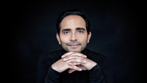 a latino man, in a black shirt that blends into the background, tents his hands below his chin as he gazes toward us