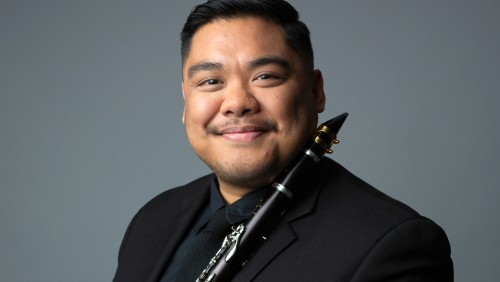 a smiling latino man with short hair and a very short facial hair holds his clarinet over his left shoulder