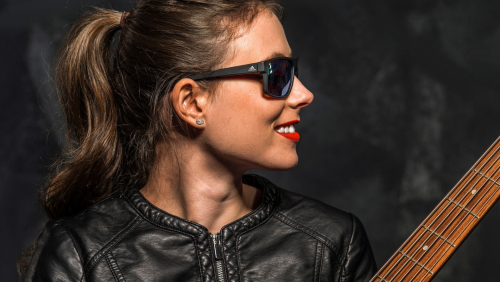 a smiling white woman with brown hair in a ponytail wears black sunglasses and a moto leather jacket. She turns toward the neck of her bass guitar.
