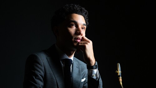 a young man in a gray suit holds a saxophone. He is starkly lit from the side and looks into the light.