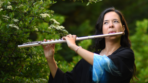 a woman in a black top plays the flute outdoors in front of green trees