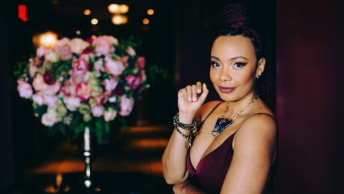 zahili zamora wears a burgundy gown, leaning on a dark wood doorway. behind her, a hallway with dramatic low light and an opulent pink floral arrangement