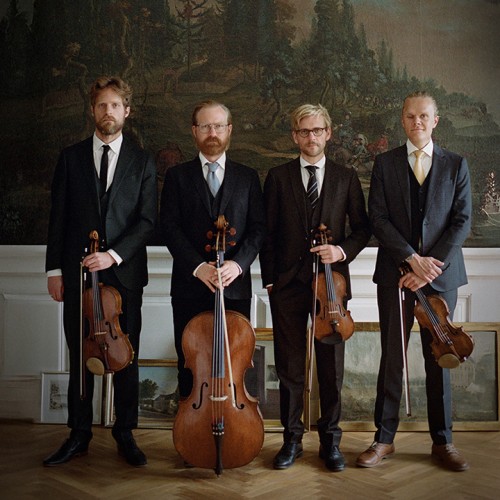 Four white men in a string quartet stand in a row in dark suits holding their instruments in front of several nature murals. 
