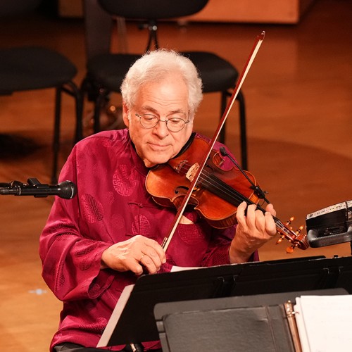An older man with white hair in a red shirt plays violin while seated in a scooter power chair. 