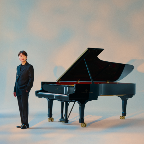 in a studio lit to resemble rosy clouds, a Korean man in his 20s stands casually a few feet away from an open grand piano.