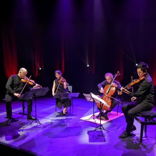 A string quartet of three men and one woman performs on a modern stage with uplighting and purple hues. 