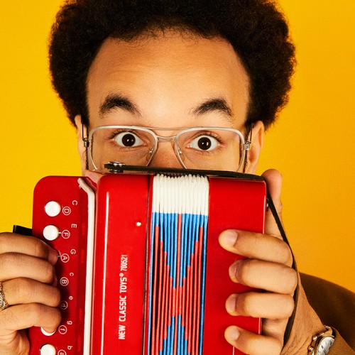 A man with a dark afro holds a toy accordion in front of his face, his eyes wide and eyebrows raised above aviator style glasses.