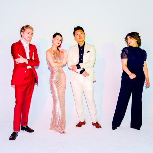 Two men and two women of a string quartet wear bold, elegant outfits, and stand before a bright backdrop under colorful light that casts multiple shadows.