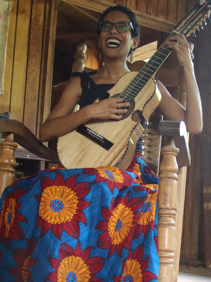 A young woman with short dark curls, glasses, and a bright floral skirt sits in a rocking chair holding a Puerto Rican cuatro. 