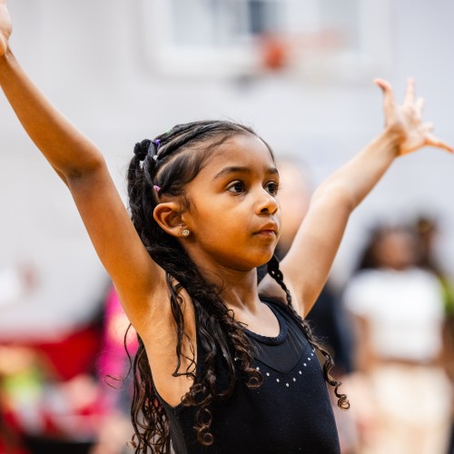a little dancer raises her arms in a wide V in a workshop class in a white gymnasium