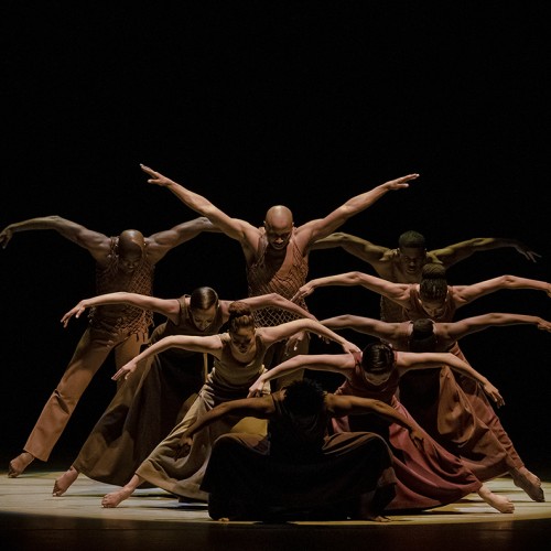 Several dancers are arranged in a series of lines, each dancer standing taller than the one in front, with arms arched beside them. 