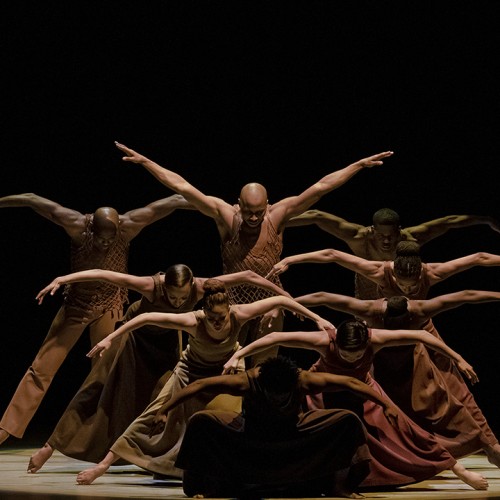 Several dancers are arranged in a series of lines, each dancer standing taller than the one in front, with arms arched beside them. 