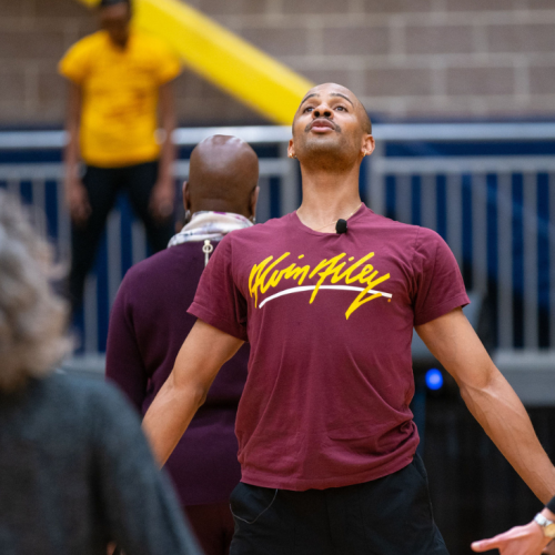 a black man in his 30s with a shaved head lifts his chest as he leads participants through a breathing or dance exercise