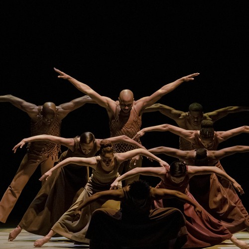 Several dancers are arranged in a series of lines, each dancer standing taller than the one in front, with arms arched beside them. 