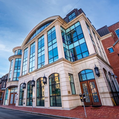 A tall stone building with many windows, surrounded by older brick buildings and a blue sky