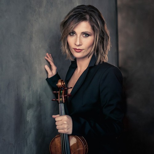 A white woman with shoulder length brown hair leans against a gray painted wall holding her violin while wearing a dark suit jacket. 